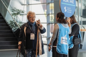 An attendee speaking to two student volunteers wearing blue bibs, one of whom is holding a lollipop sign. 